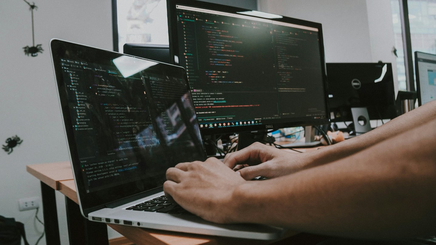 Hands typing code on a laptop connected to a large monitor.
